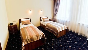 A hotel room with two single beds, a bedside table with a lamp, an air conditioning unit, and a window with sheer curtains.