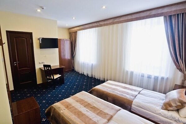 A hotel room with two beds, a desk, a chair, a TV, and large windows with curtains.
