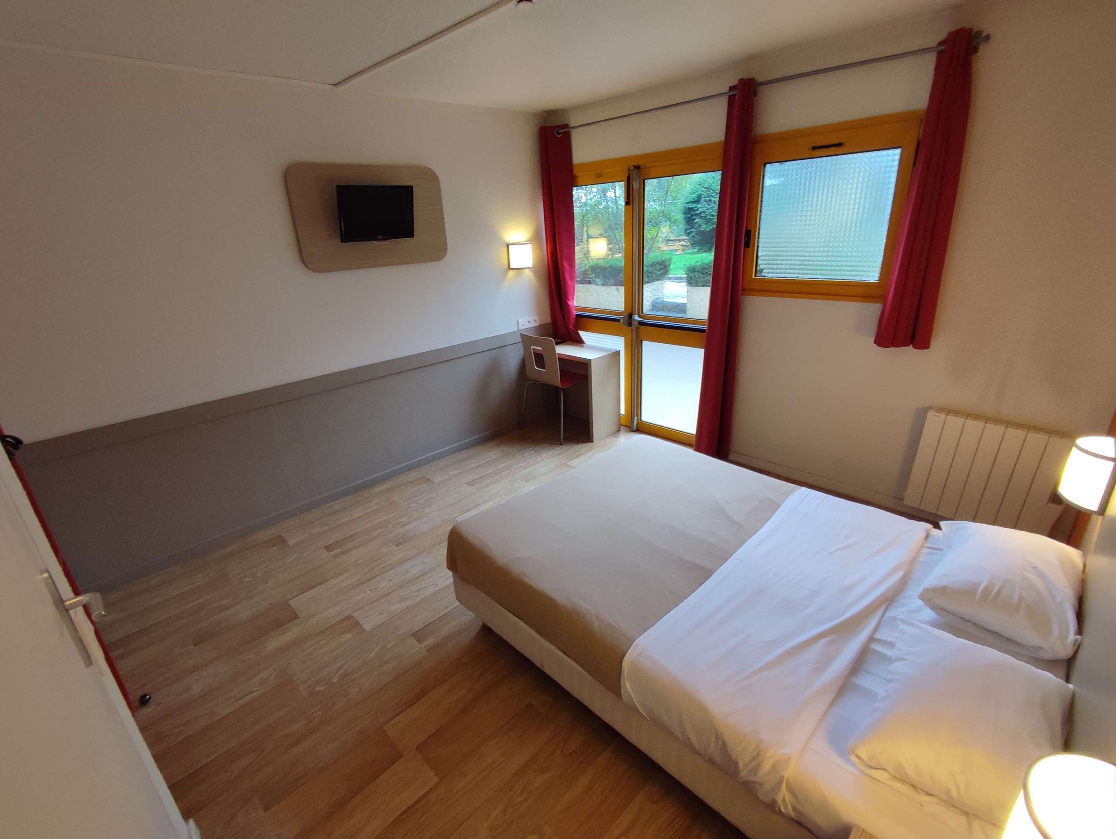 Une chambre d’hôtel avec un lit, un bureau, une chaise, une télévision et une fenêtre avec des rideaux.
