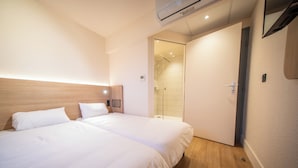 A hotel room with two beds, a wooden headboard, a shower area, and a door leading to another room.