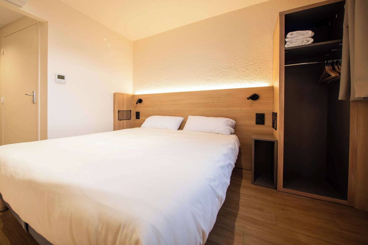 Una habitación de hotel moderna con una cama grande, un cabecero de madera, un armario empotrado y una mesita de noche pequeña.