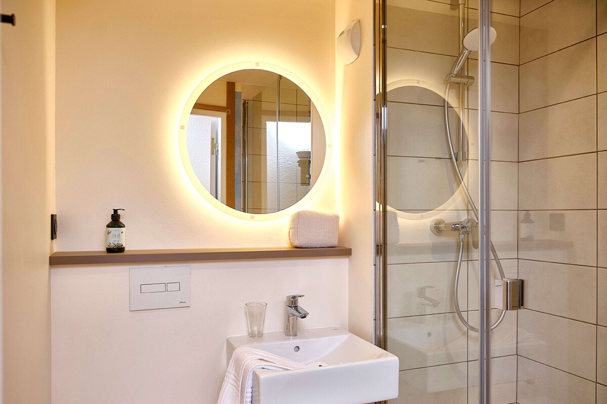 Een moderne badkamer met een douchecabine van glas, een wit wastafelmeubel, een spiegel met een rond verlichte omlijsting en een klein plankje met een fles en een handdoek.