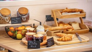 A breakfast buffet with jars of jam, a bowl of fruit, and a wooden serving board with pastries. - Premiere Classe Annecy Cran-Gevrier (Annecy)