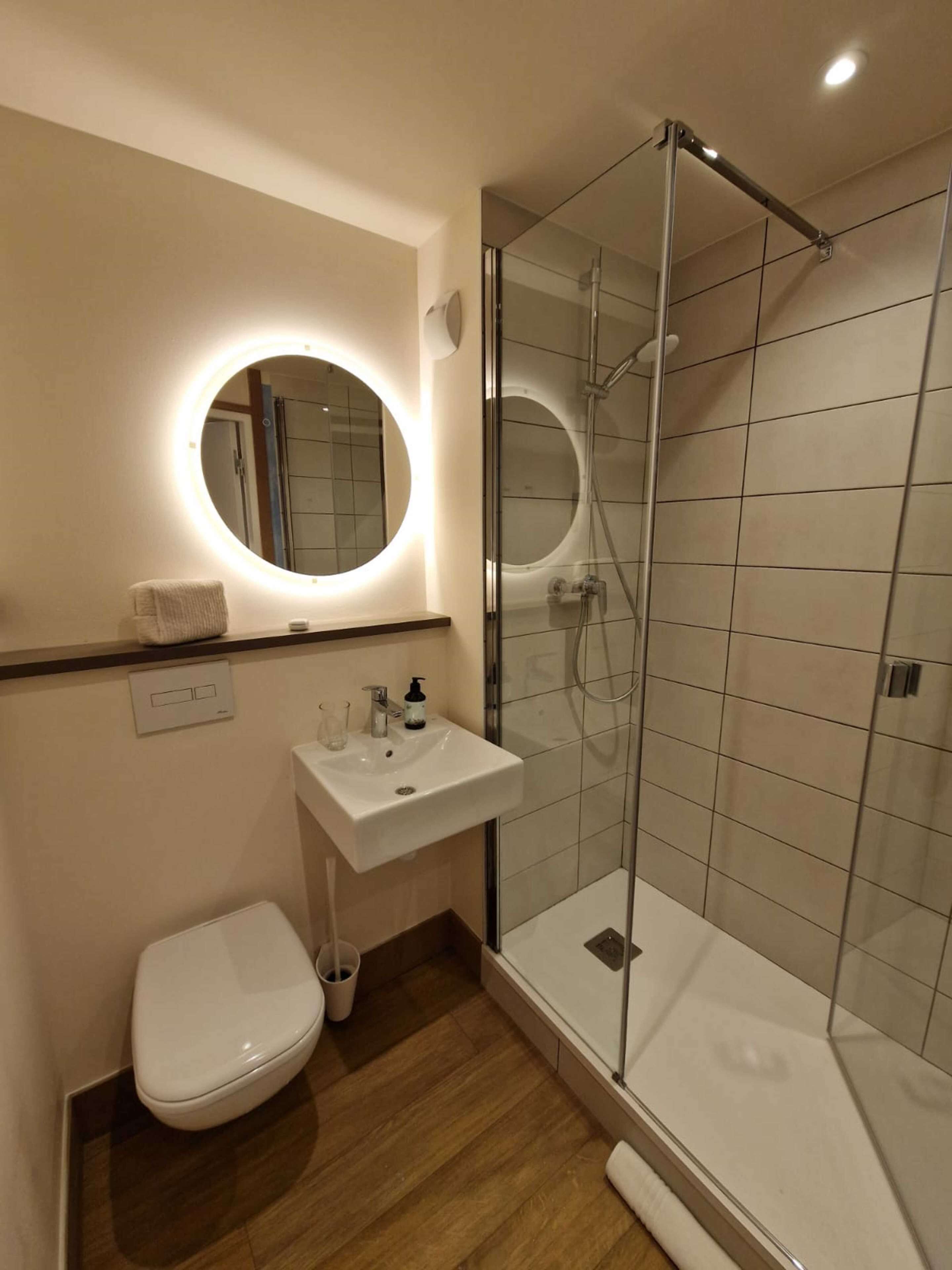 A modern bathroom with a glass-enclosed shower, a round mirror with built-in lighting, a white sink, and a toilet.