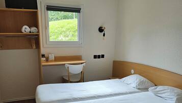 A bedroom with two beds, a desk, and a window with a view of greenery.