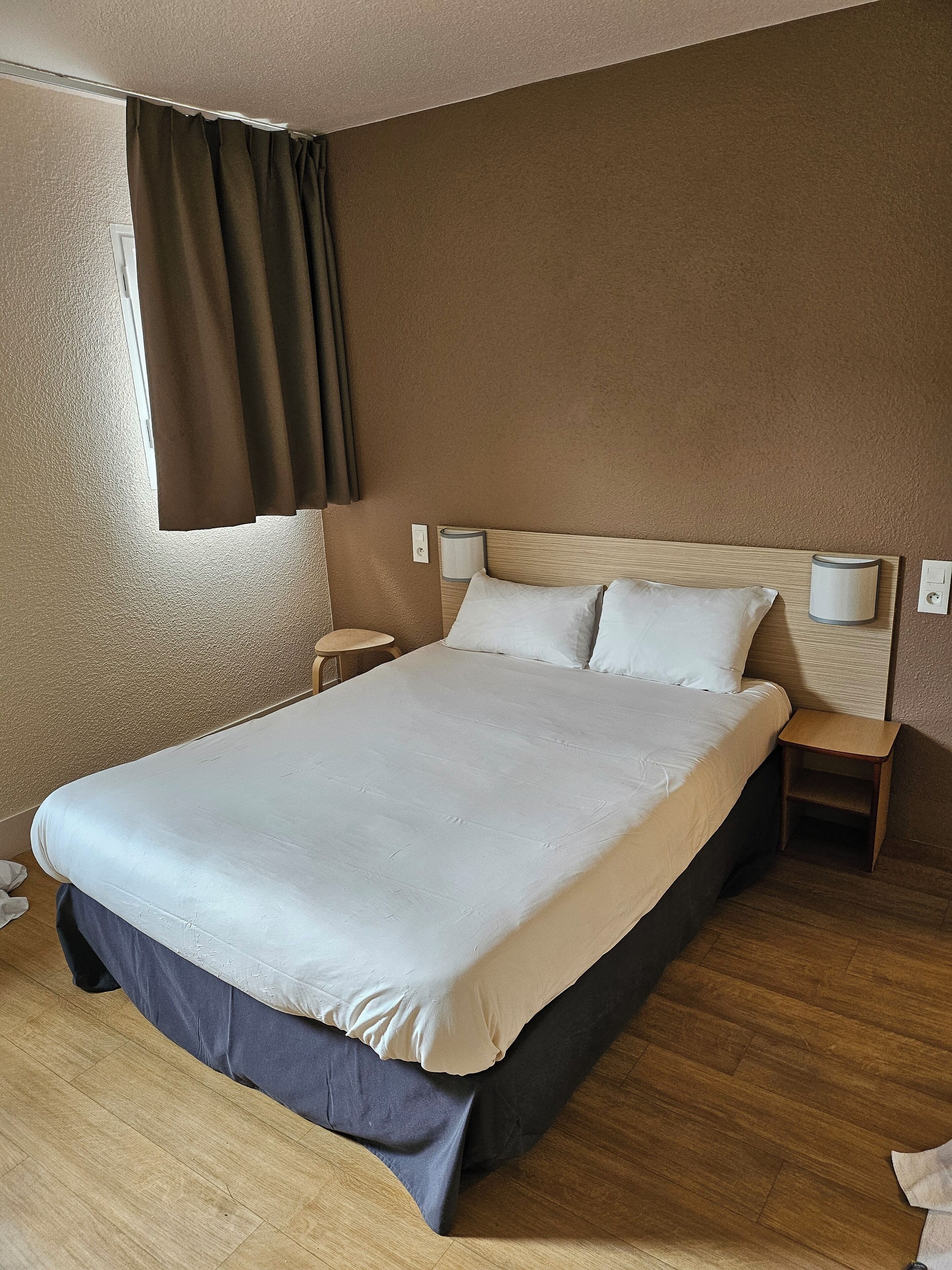 A hotel room with a large bed, two bedside tables, a nightstand, a window with curtains, and a wooden floor.