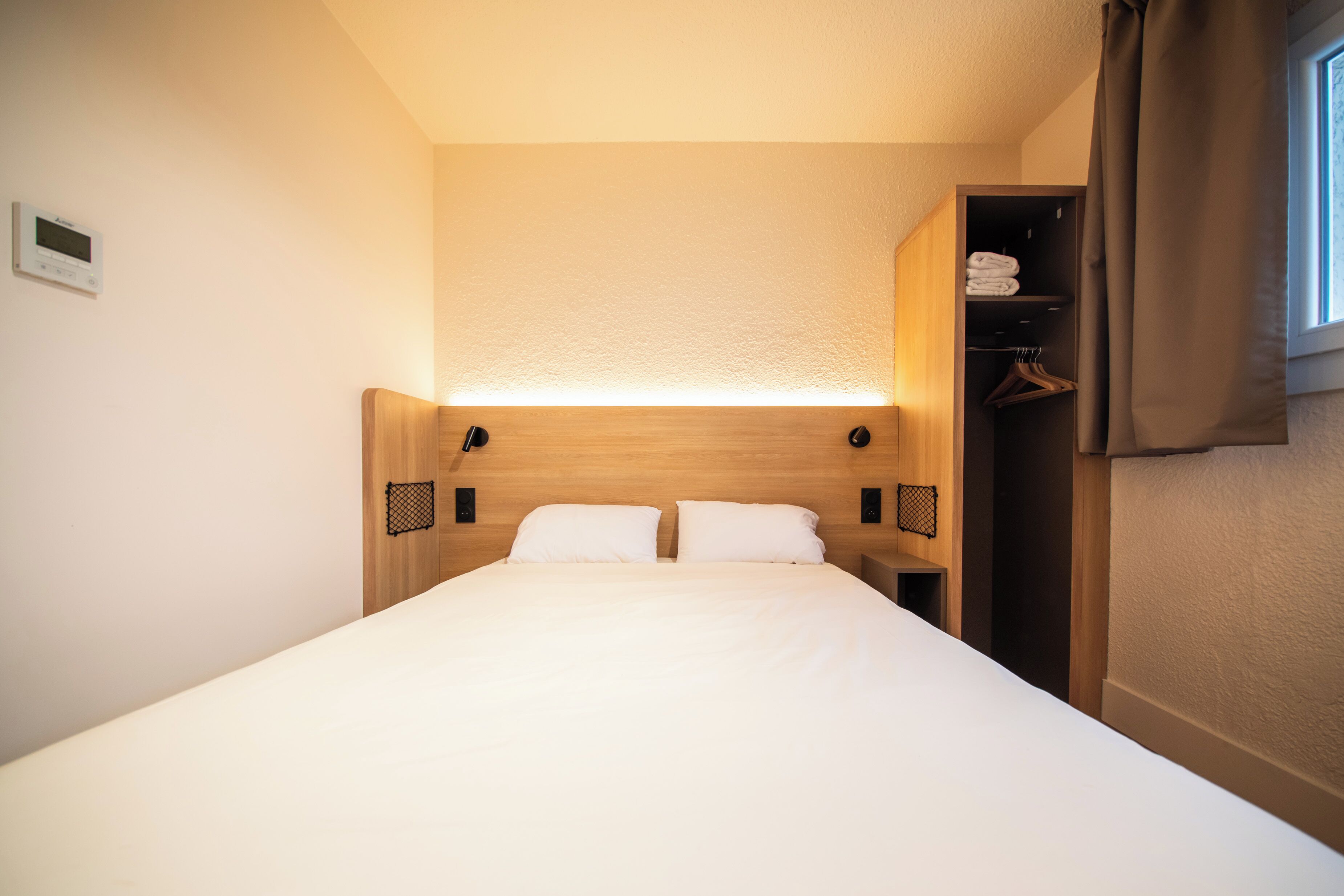 A neatly made bed with a wooden headboard, a bedside table, and a hanging closet with clothes.