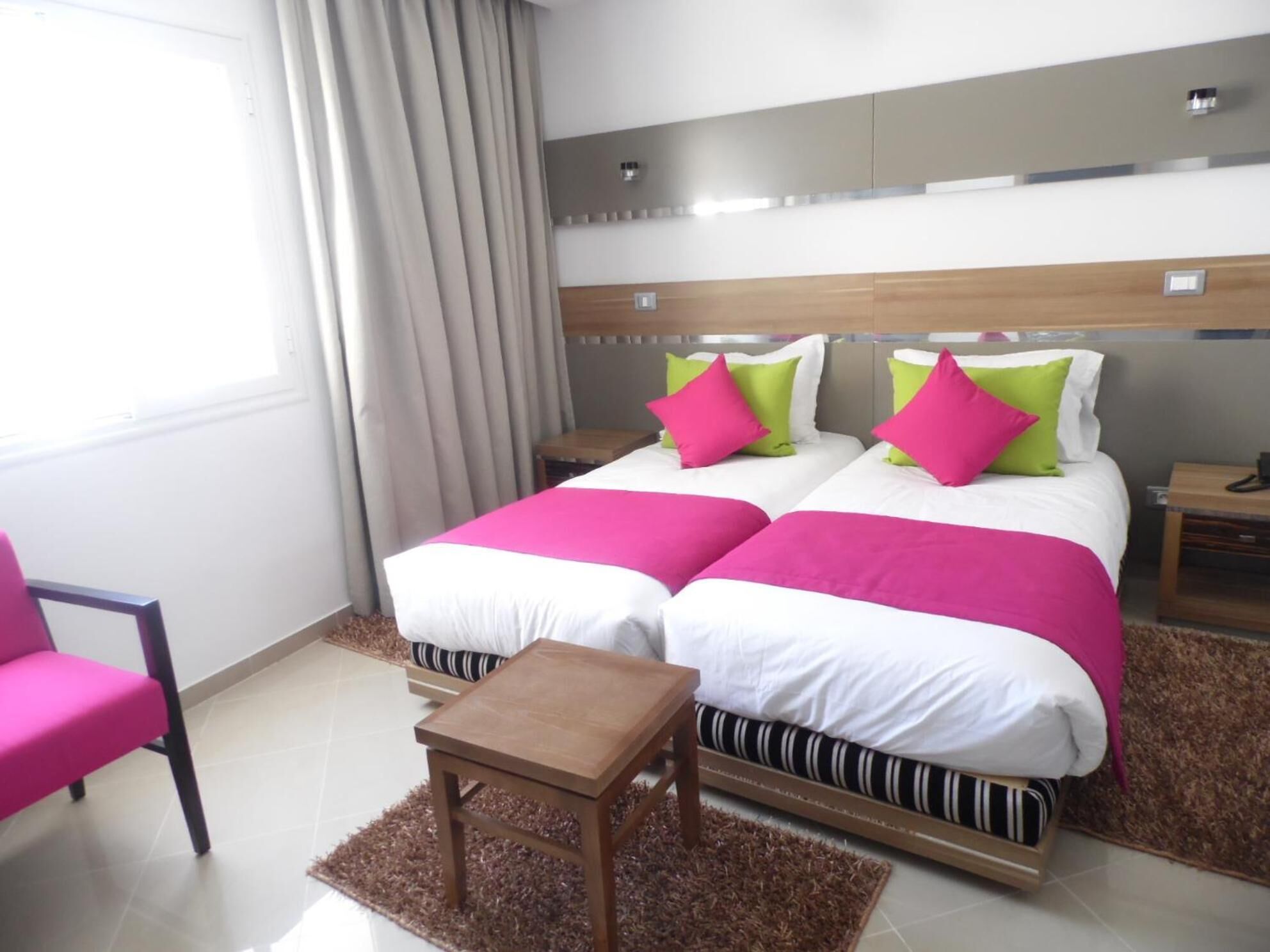 Habitación de hotel con dos camas, una mesita de madera, una silla y un ventanal con cortinas.