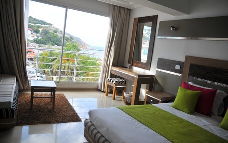 A hotel room with a bed, a desk, a chair, a small table, and a view of a coastal area.