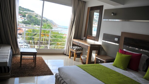 A hotel room with a bed, a desk, a chair, a small table, and a view of a coastal area. - Hotel Marina Prestige Tabarka (Tabarka)