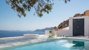 A clear blue pool with a view of the sea and mountains, a white building with a dark door, and a small plant on the terrace. - Pina Caldera Residence (Santorini)