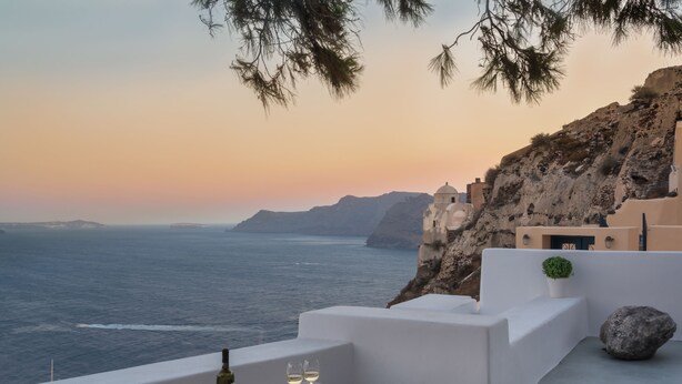 A seaside terrace with a view of the ocean and cliffs, featuring a table with a bottle and glasses, and a plate of fruit. - Pina Caldera Residence (Santorini)