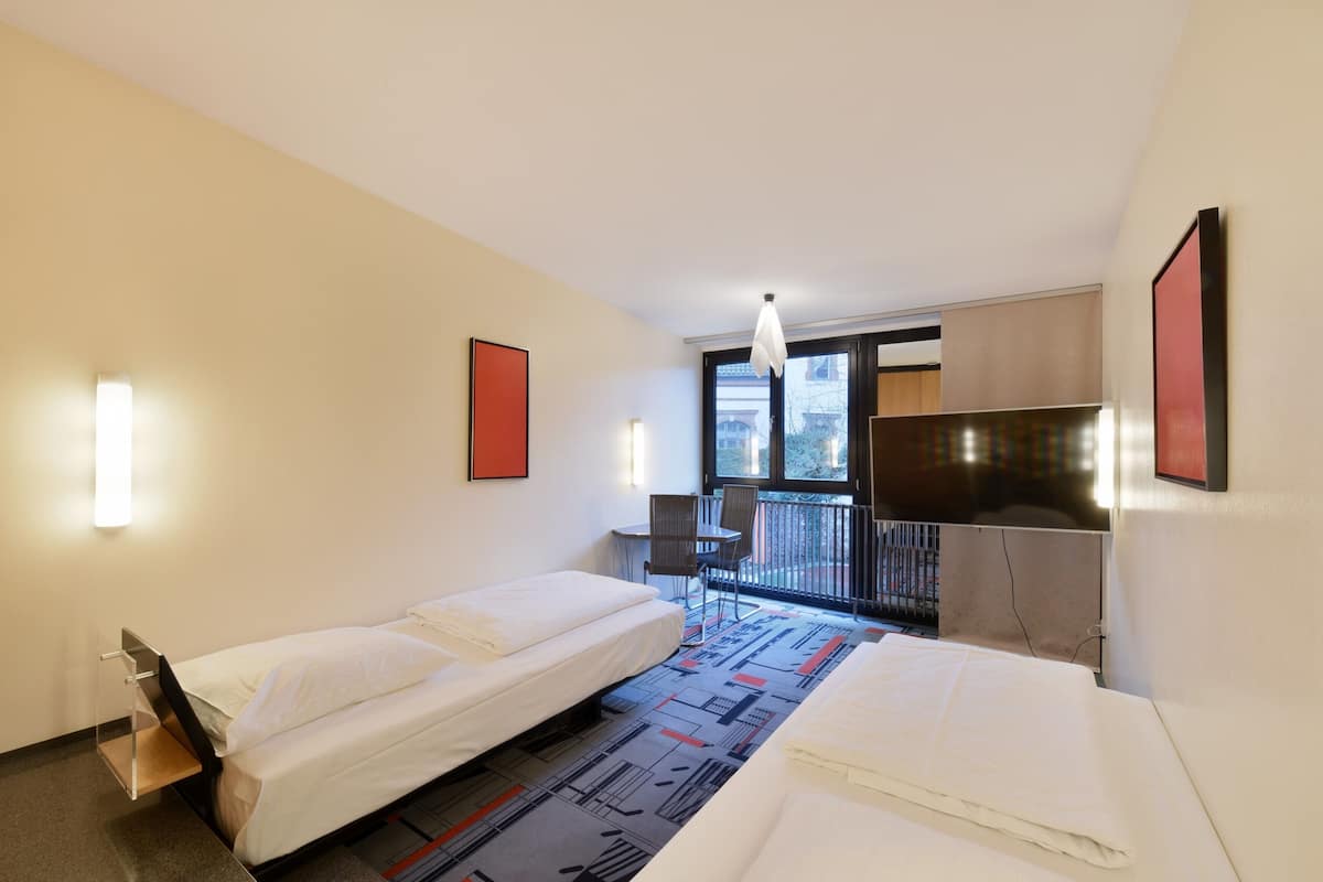 Hotellihuone, jossa on kaksi erillistä sänkyä, pieni pöytä, tuoleja, parveke ja suuri ikkuna.