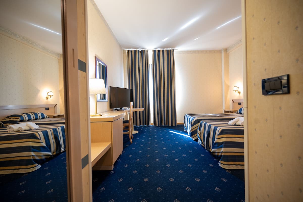 Ein Hotelzimmer mit zwei Betten, einem Schreibtisch mit Fernseher und einem großen Fenster mit gestreifteren Vorhängen.