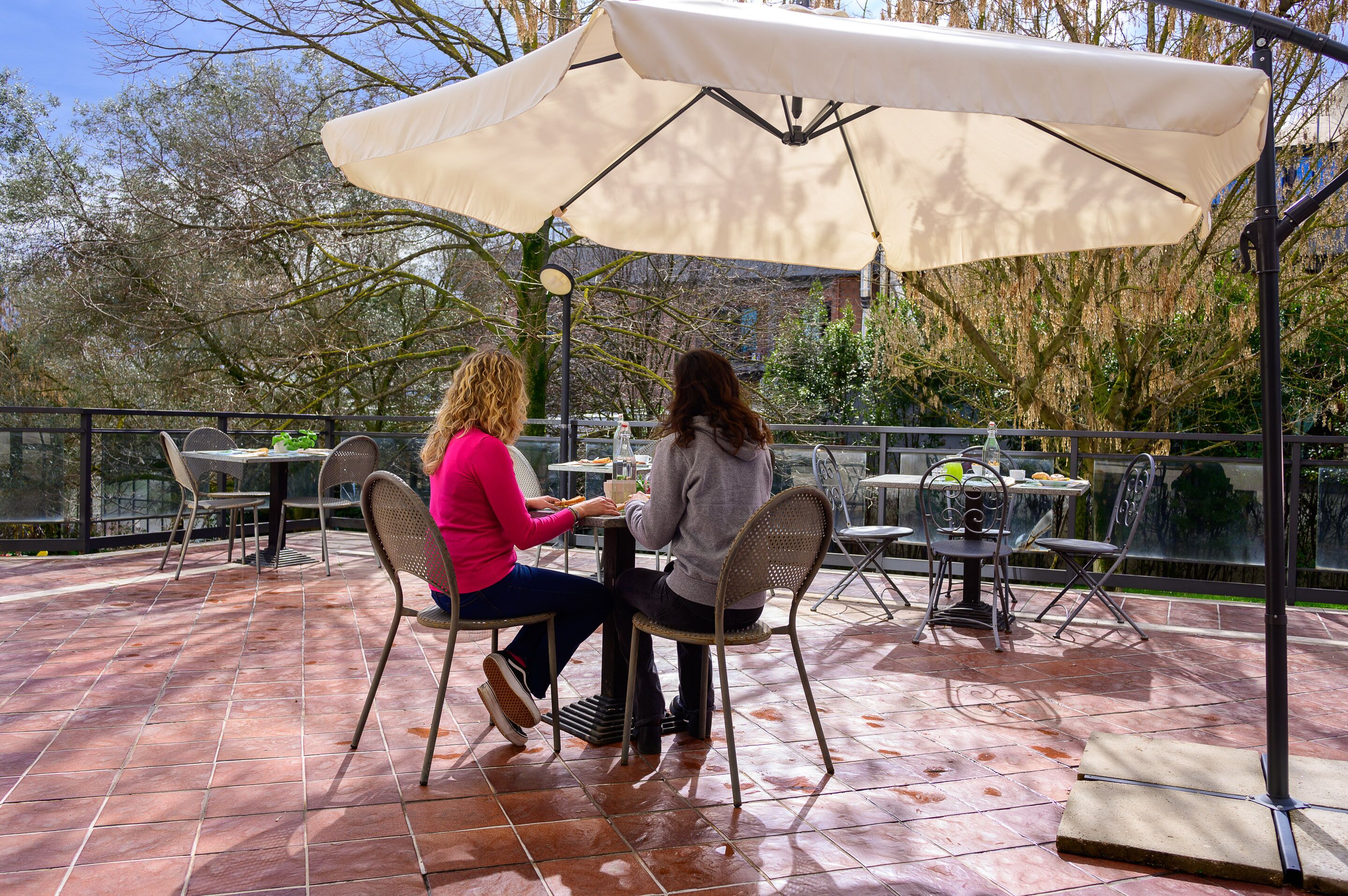 Dos personas sentadas a una mesa bajo un paraguas grande en un patio al aire libre con suelo de baldosas y sillas de metal.