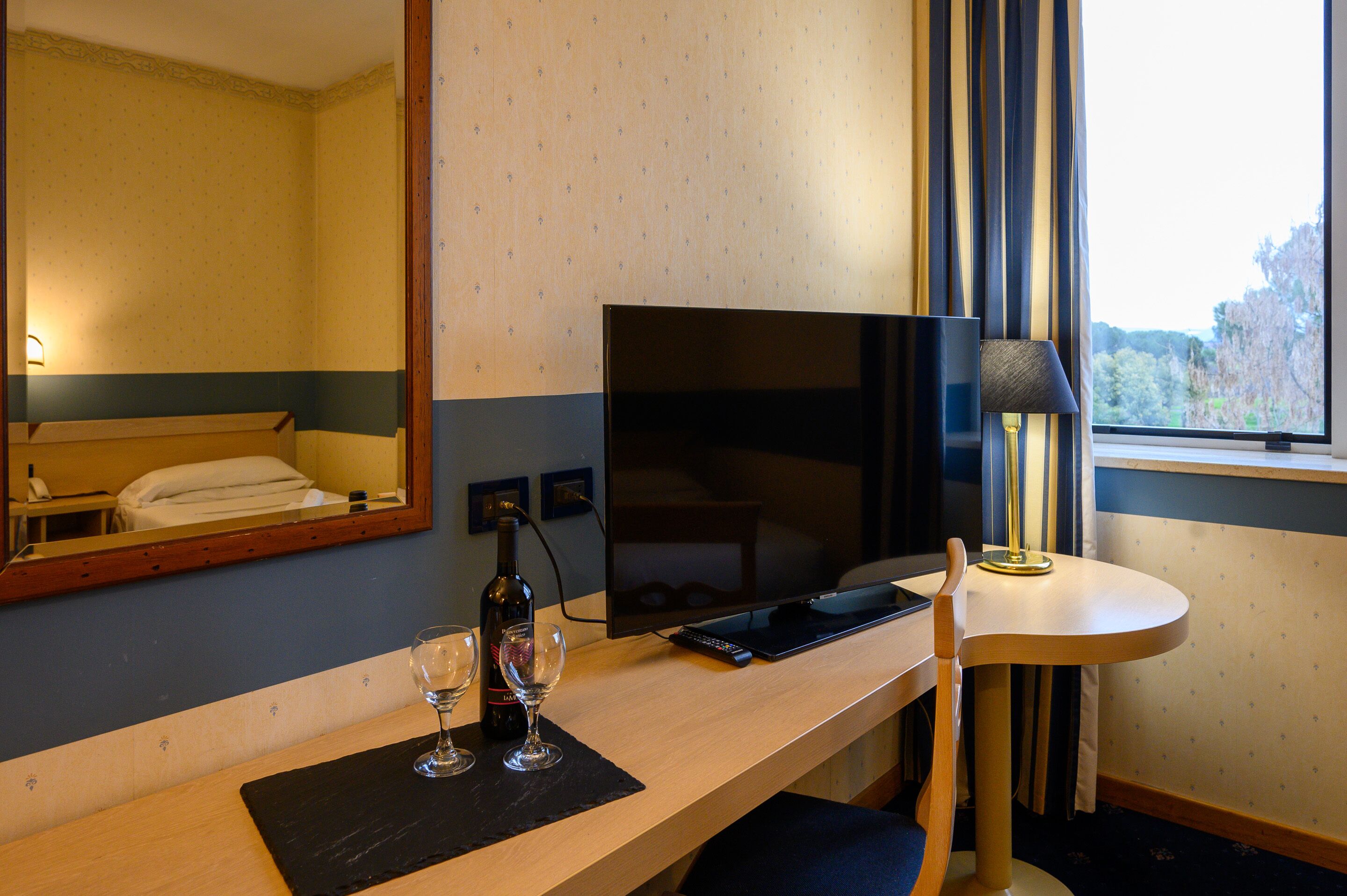 Una habitación de hotel con cama, escritorio con televisor, lámpara y ventana con cortinas.