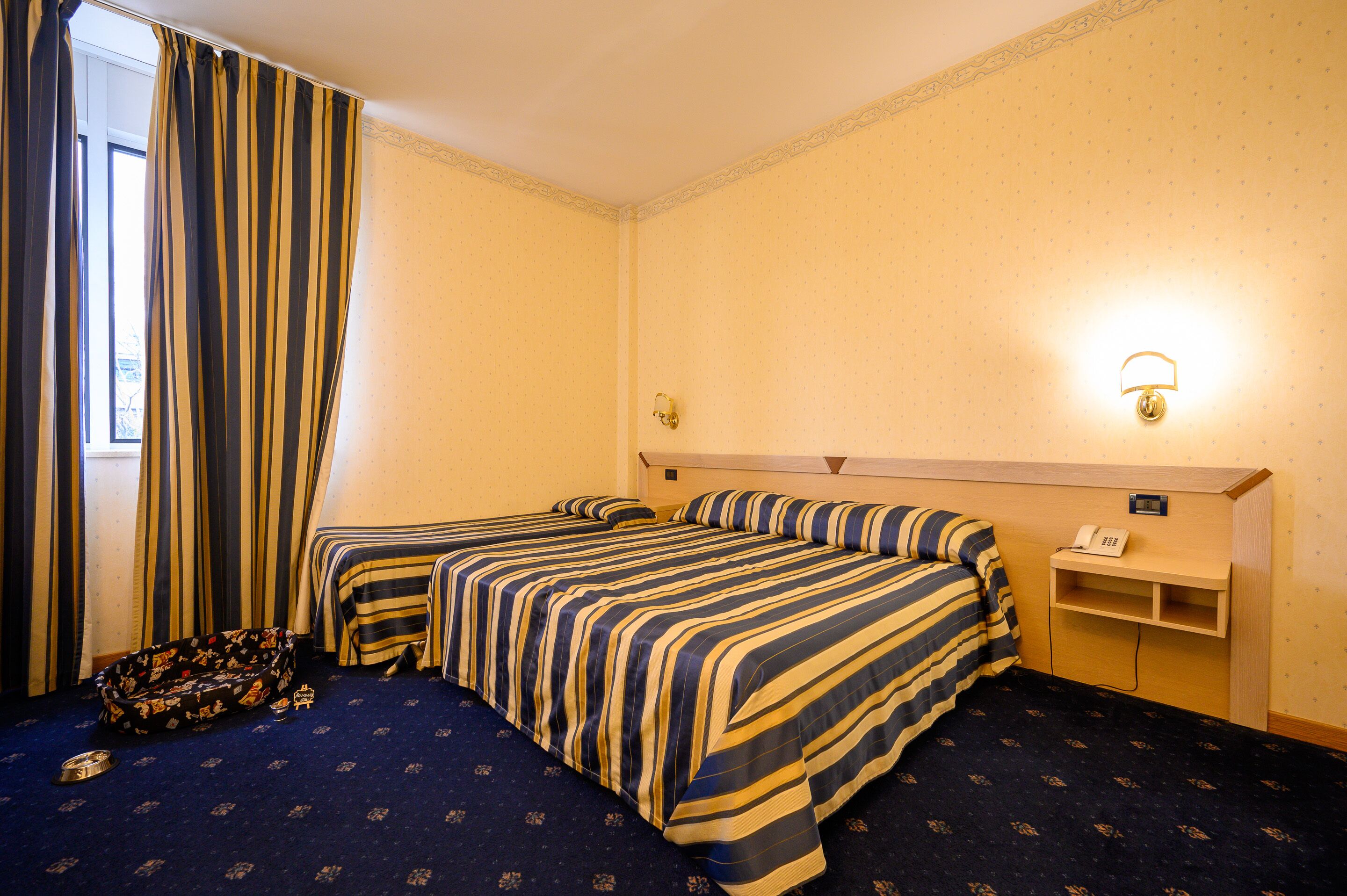 A hotel room with two beds, a small desk with a telephone, and a window with curtains.