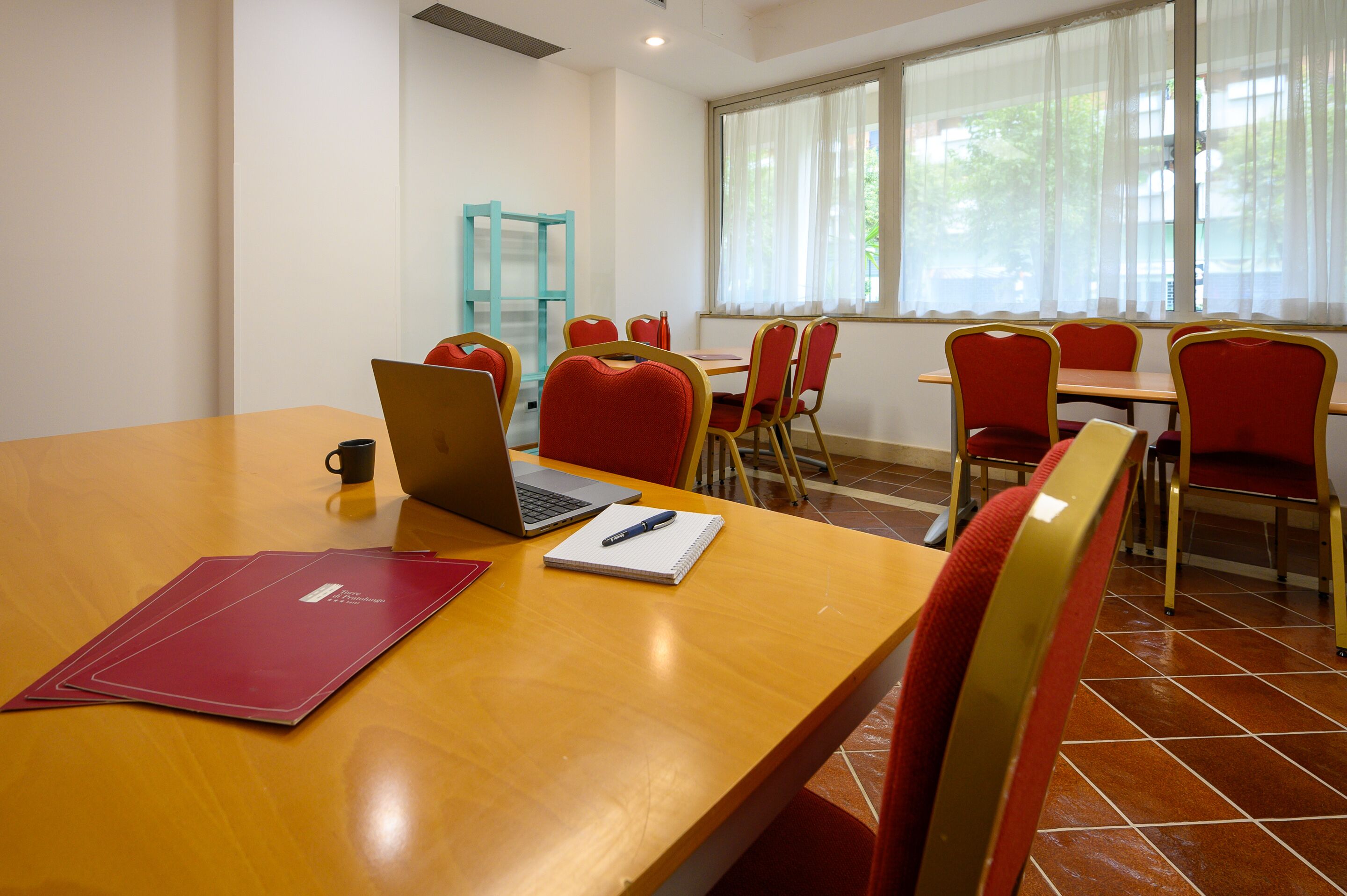 A conference room with a wooden table, red chairs, a laptop, a notebook, and a pen.