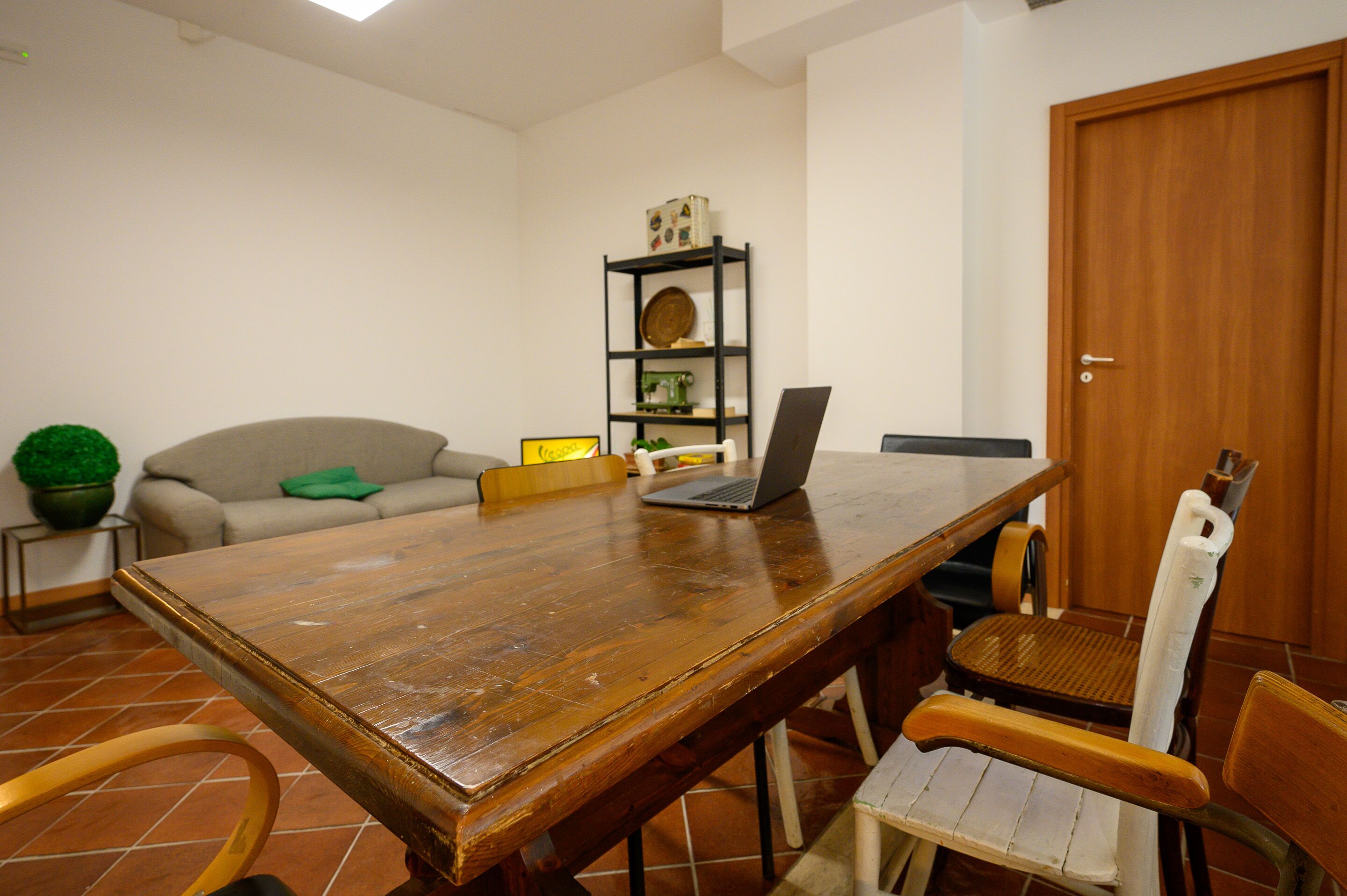 A wooden dining table with a laptop, a chair, and a bookshelf in a room with a tiled floor and a door.