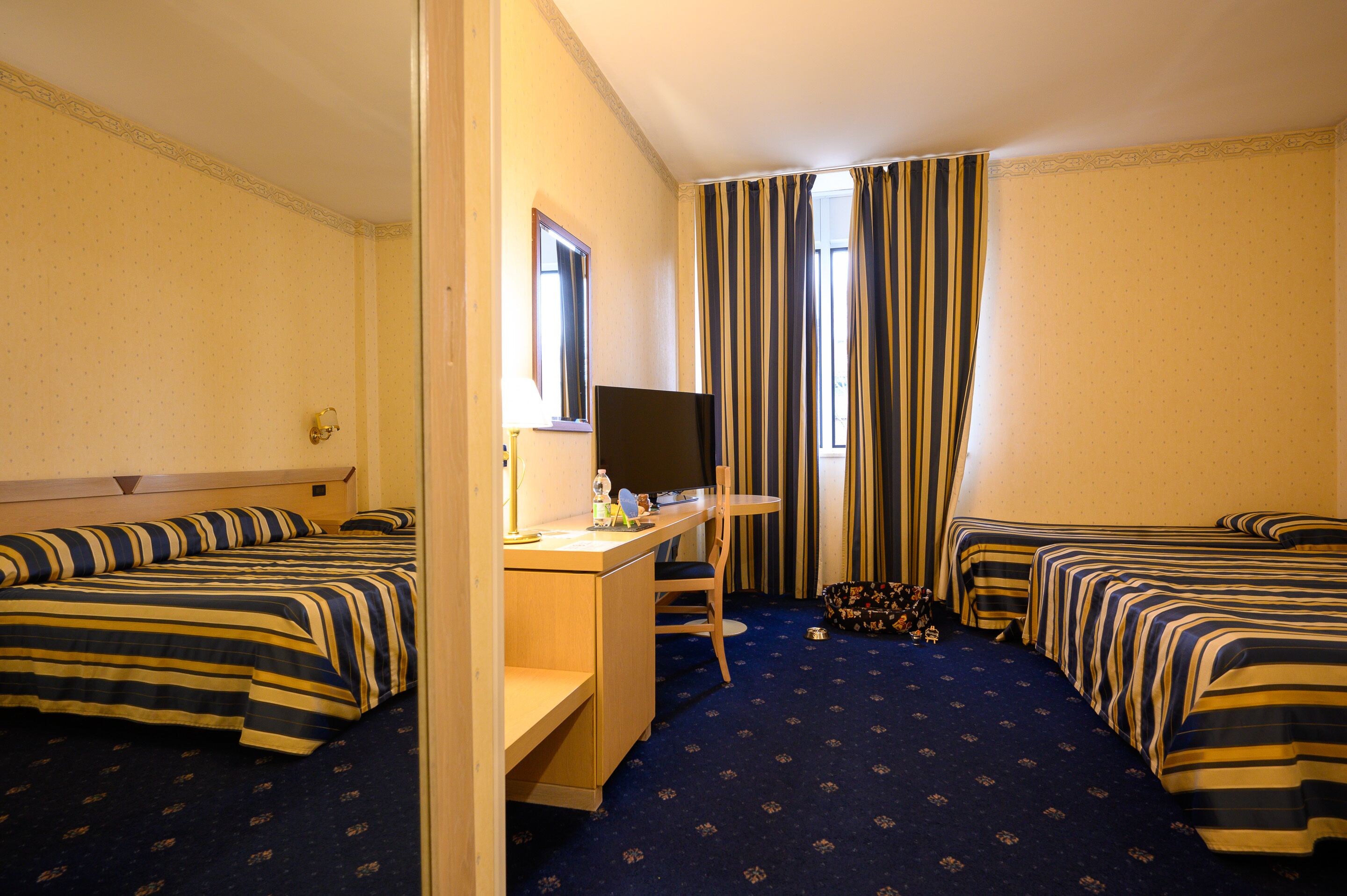 Habitación de hotel con dos camas, un escritorio, una silla, un televisor y una ventana con cortinas a rayas.