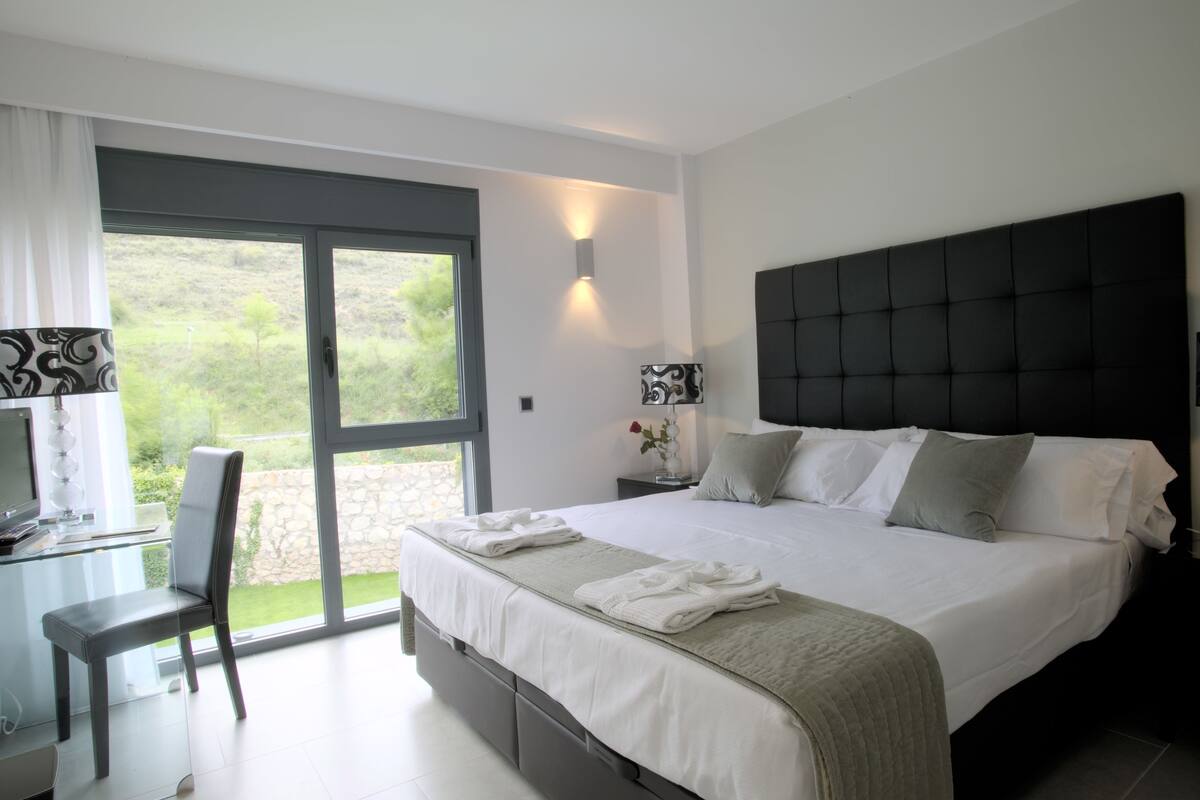 A modern bedroom with a large bed, a desk with a chair, a TV, and a view of greenery through the sliding glass door.