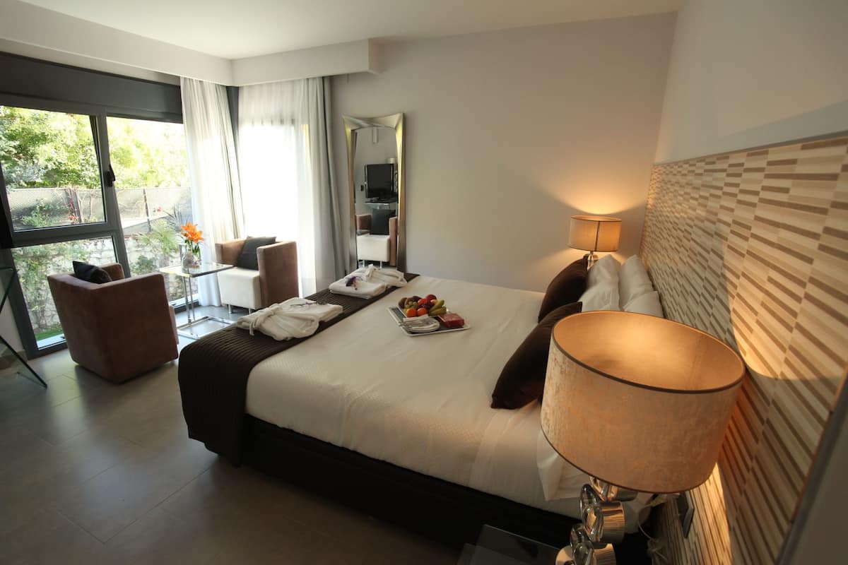A modern hotel room with a large bed, a sitting area with a chair and table, a large window, and a mirror.