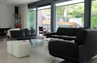 Una sala de estar moderna con un sofá de cuero negro, un reposapiés blanco y una mesa de centro de vidrio. Una ventana grande ofrece vistas al exterior.