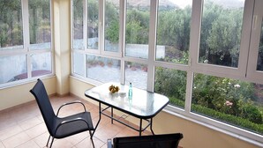 Une pièce avec une table en verre, deux chaises et de grandes fenêtres donnant sur un jardin et des collines.