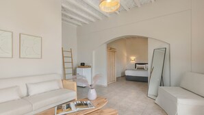 Una sala de estar moderna con un sofá, una mesa de centro y una zona de lectura. Se ve una escalera, una cesta y un jarrón con plumas. Un arco de entrada conduce a otra habitación con una cama y un espejo.