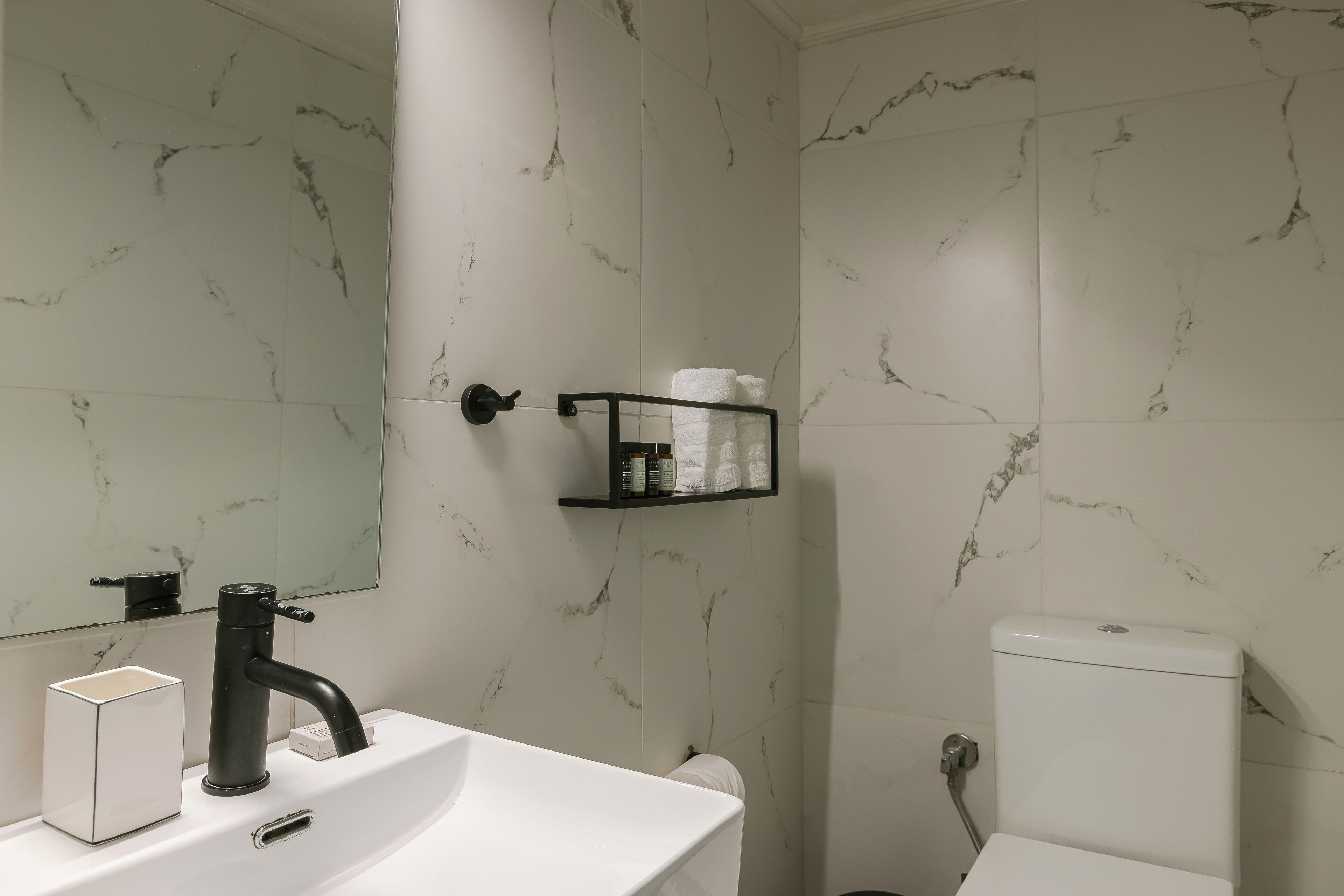 A bathroom with a white sink, black faucet, marble-patterned walls, a toilet, and a shelf with toiletries.