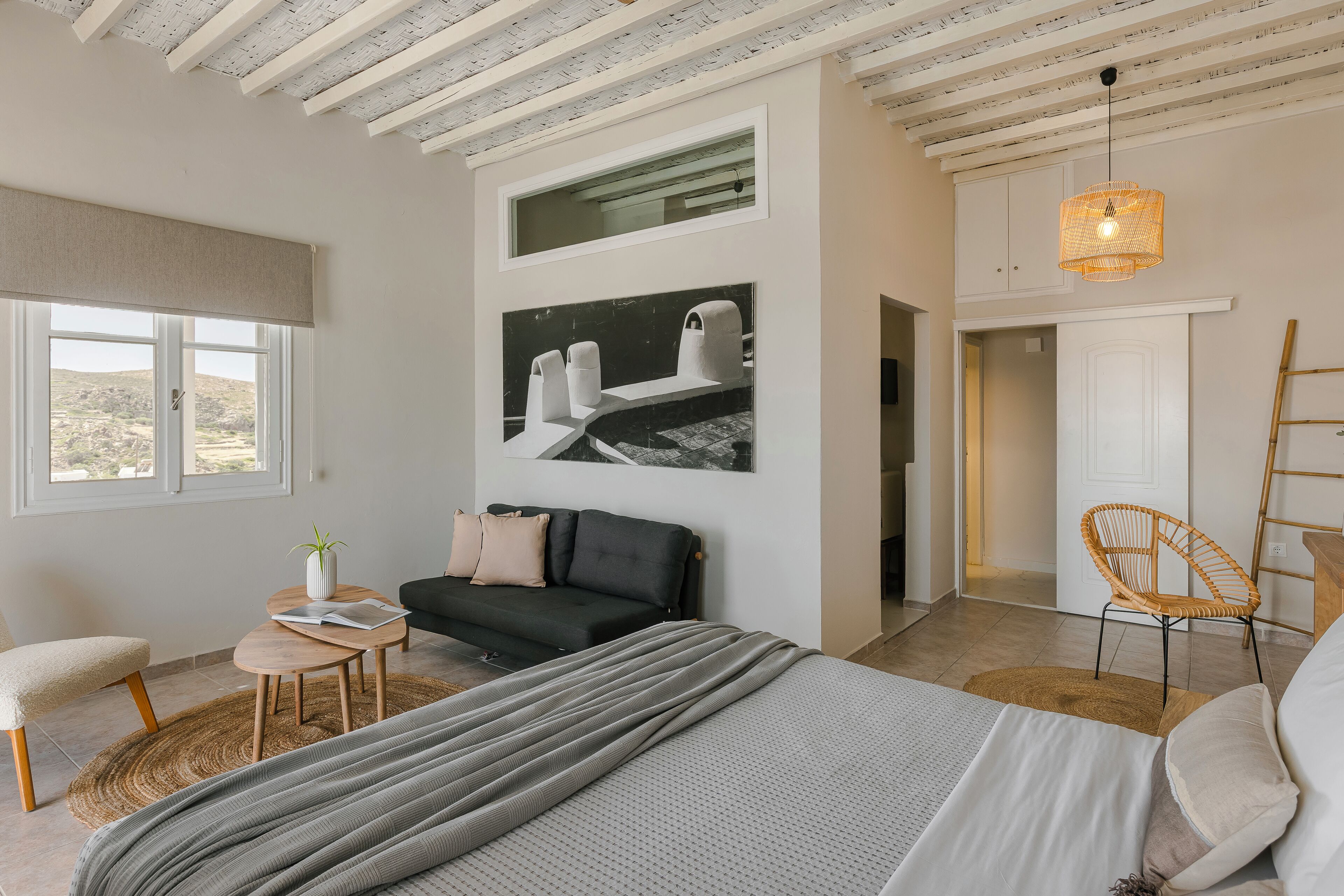 A modern bedroom with a bed, a sofa, a dining table, and a chair. There is a window with a view of the outdoors.