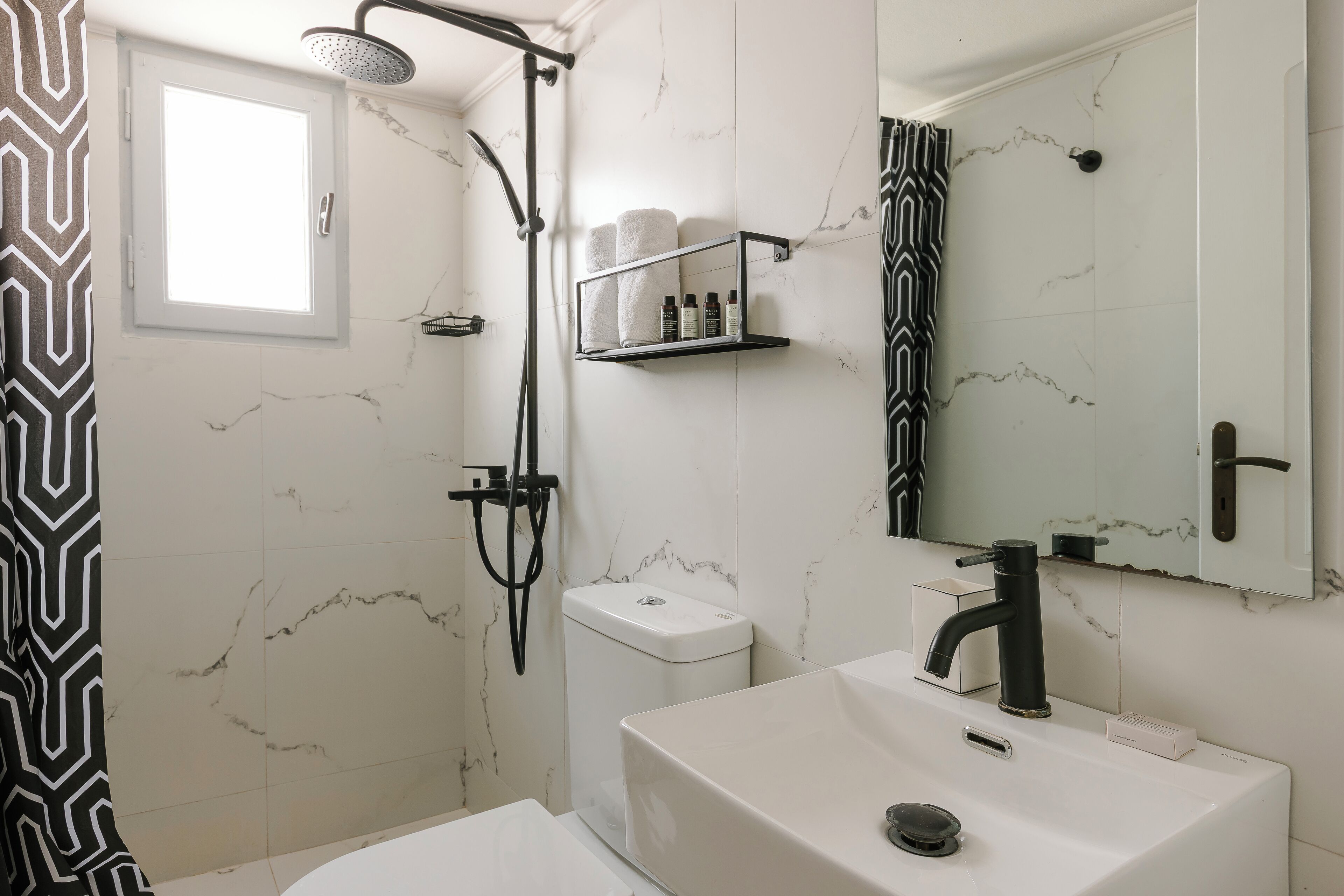 Un baño con lavamanos blanco, grifo negro y ducha con cabezal de ducha manual. Hay una repisa con artículos de aseo y un perchero sobre el lavamanos.