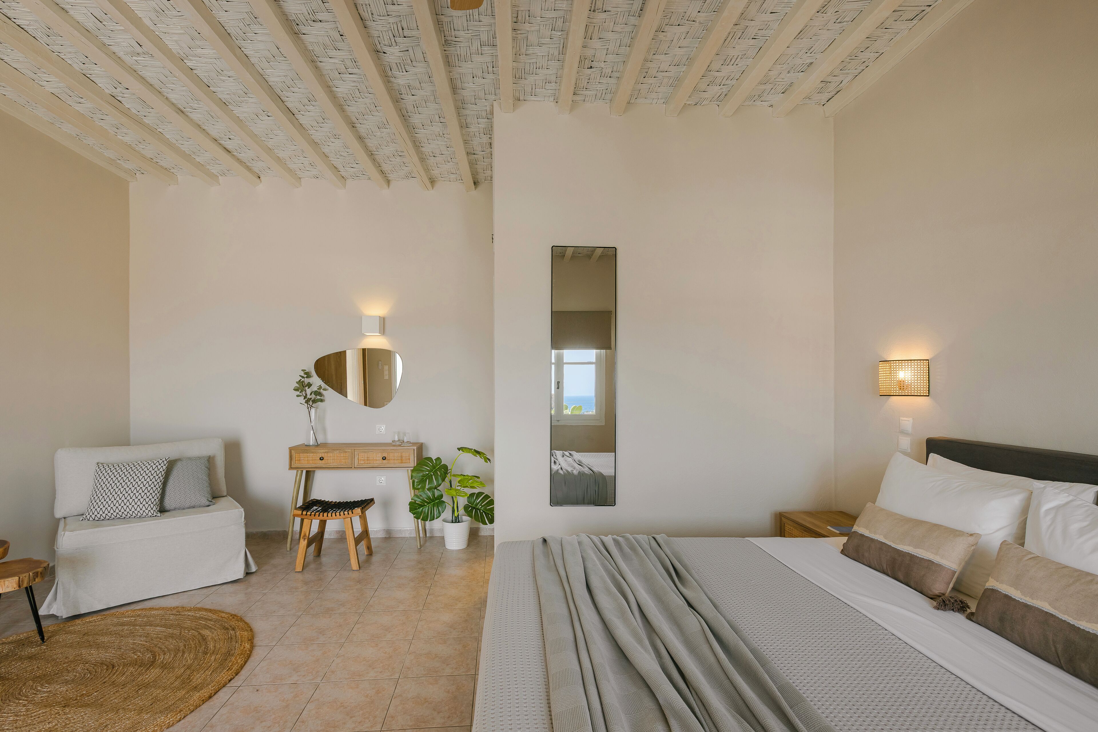 Un dormitorio con cama, escritorio, silla, una mesita, una planta en maceta y un espejo en la pared.