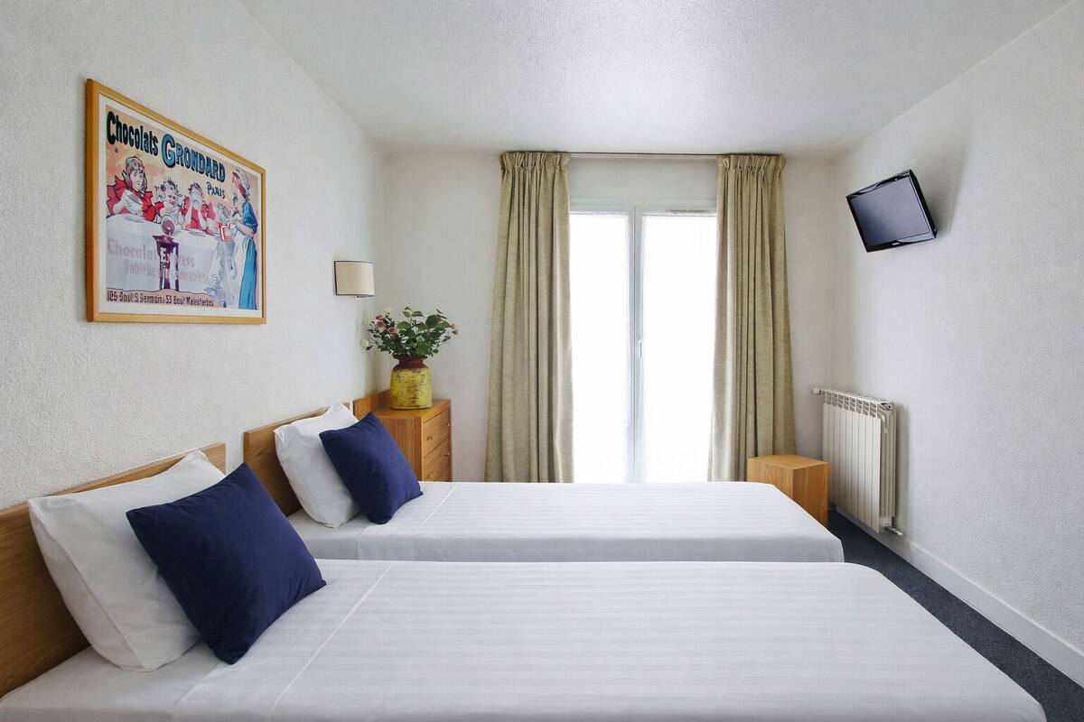 Ein Hotelzimmer mit zwei Betten, einem an der Wand befestigten Fernseher, einem Fenster mit Vorhängen, einem Heizkörper und einem Poster an der Wand.