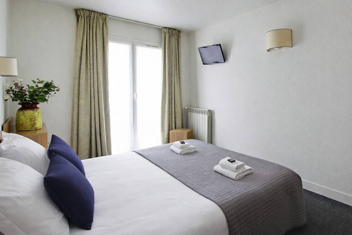 Ein Hotelzimmer mit einem Bett, zwei Kissen, einem Nachttisch mit Lampe, einer Vase mit Blumen, einem an der Wand befestigten Fernseher und einem Heizkörper.