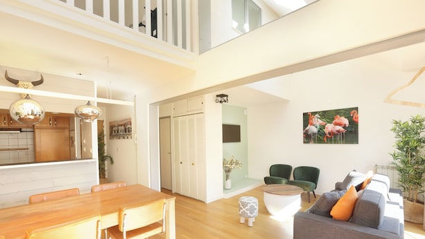A spacious living area with a wooden dining table, a gray sofa with orange cushions, and a painting of flamingos on the wall. -  Les Lilas Serviced Apartments (Les Lilas)