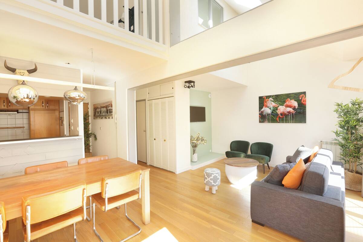Ein geräumiger Wohnbereich mit einem Holzessttisch, einem grauen Sofa mit orangefarbenen Kissen und einem Flamingo-Bild an der Wand.