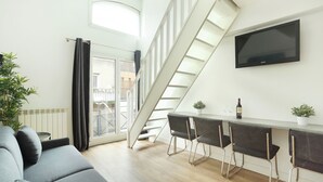 A modern living space with a sofa, a dining table with chairs, a TV mounted on the wall, and a staircase leading to an upper level. -  Les Lilas Serviced Apartments (Les Lilas)