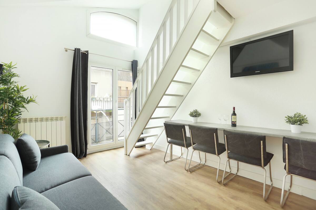 Ein moderner Wohnraum mit Sofa, einem Esstisch mit Stühlen, einem an der Wand montierten Fernseher und einer Treppe zu einer oberen Ebene.