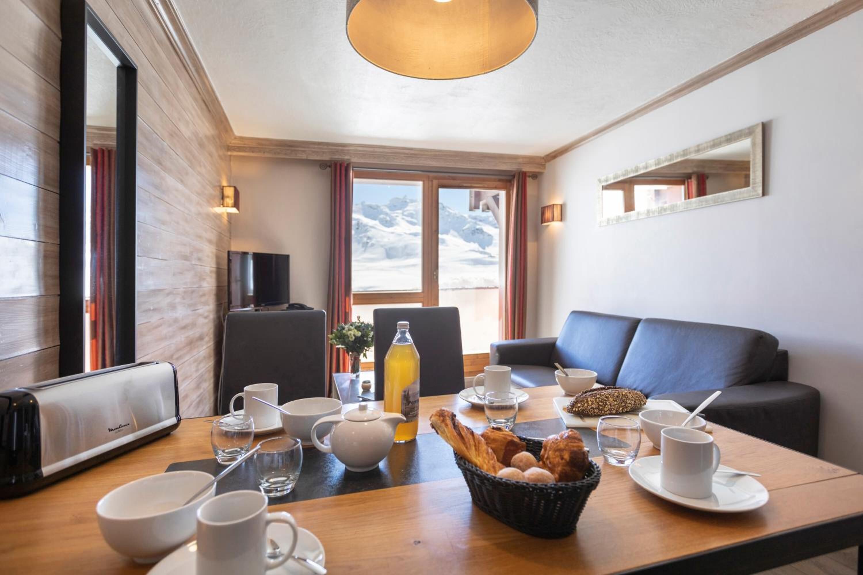 Un comedor con mesa de madera dispuesta para el desayuno, incluyendo cuencos, tazas y una cesta de pan.
