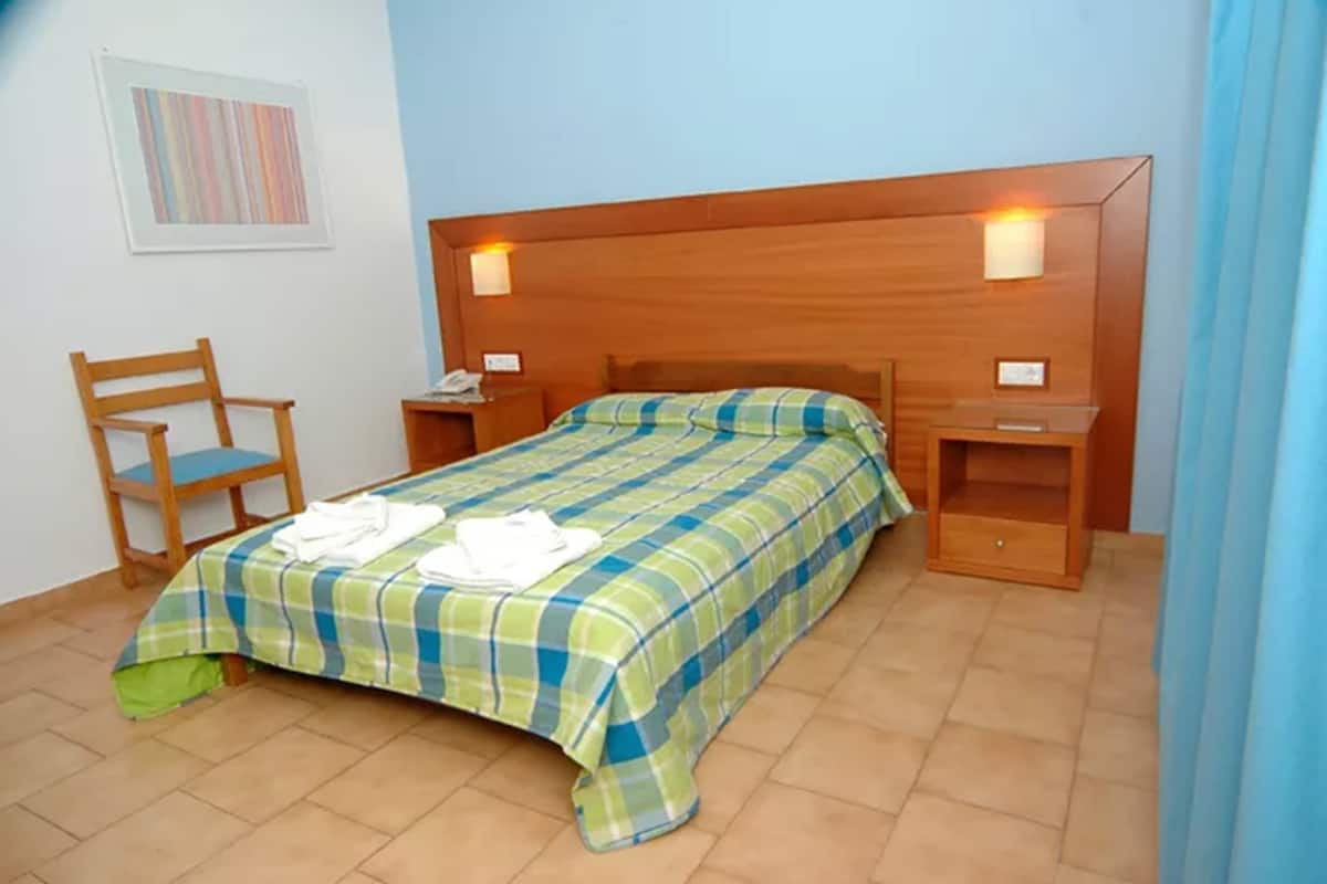 Cama bem arrumada com colcha xadrez, cabeceira de madeira, duas mesas de cabeceira com abajures e uma cadeira.