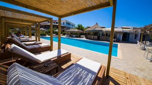 A poolside area with wooden loungers, a clear blue pool, and a thatched-roof structure in the background. - Navarone (Pylos-Nestoras)