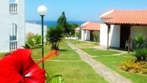 A pathway leading to a row of white buildings with red-tiled roofs, a clear blue sky, and a red flower in the foreground. - Navarone (Pylos-Nestoras)
