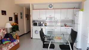 A compact living space with a kitchenette, a glass dining table with four chairs, a sofa with colorful cushions, and a washing machine. - Apartamentos Surfing 3000 (Peniscola)