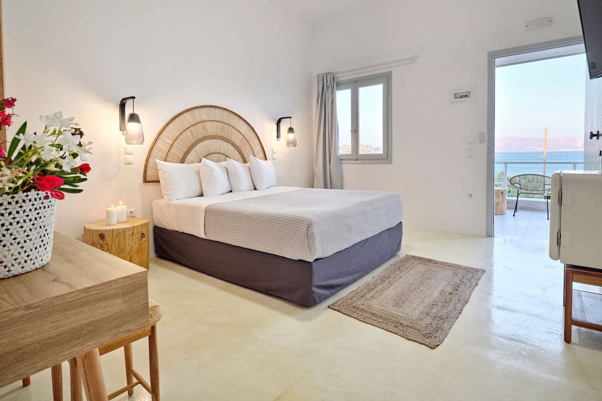 Een slaapkamer met een bed, nachtkastje, lamp en uitzicht op de zee door een raam.