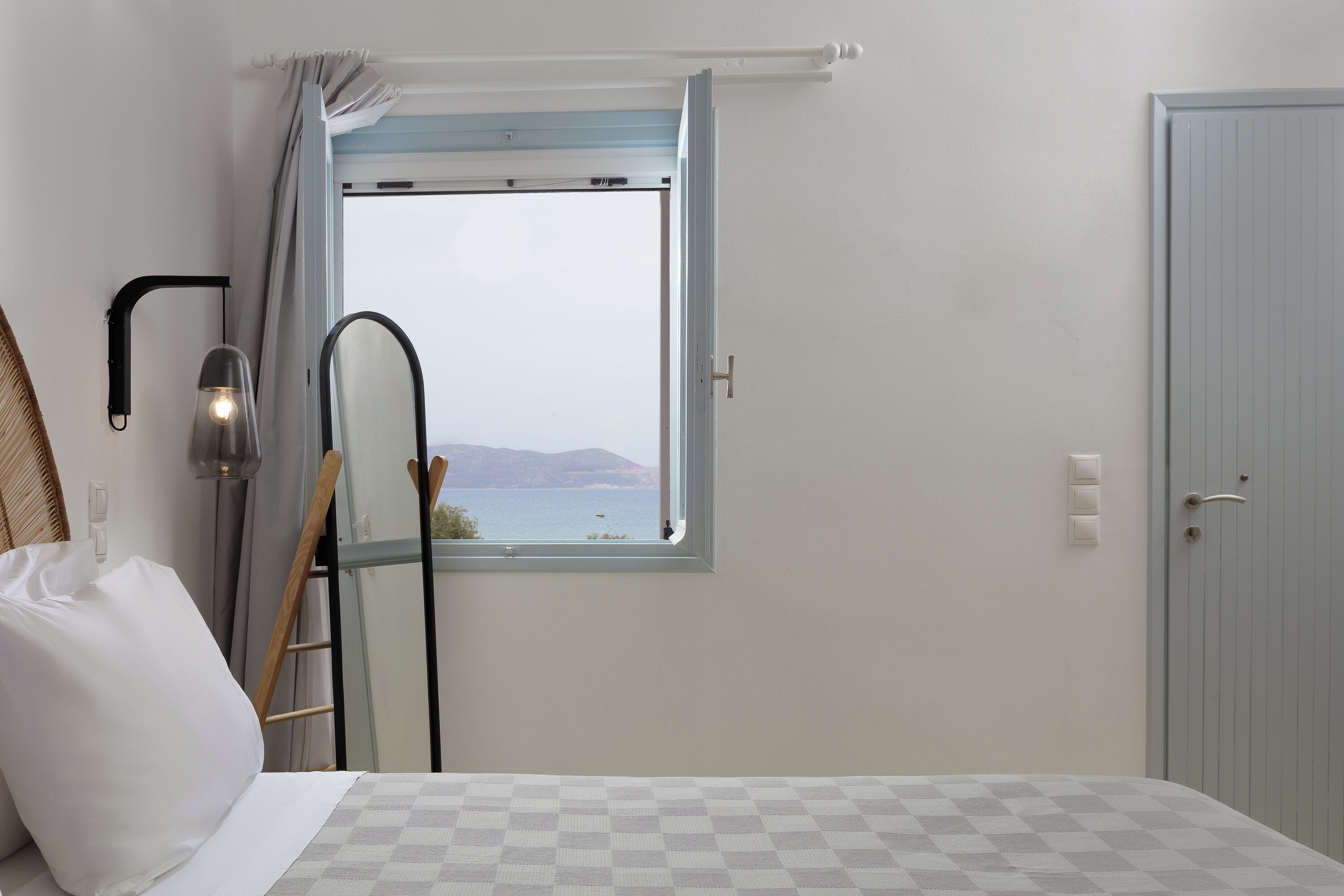 A bedroom with a checkered bedspread, a view of the sea through a window, a ladder leaning against the wall, and a door to the right.