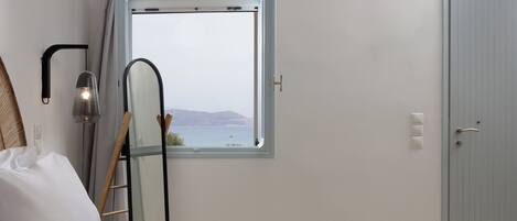A bedroom with a checkered bedspread, a view of the sea through a window, a ladder leaning against the wall, and a door to the right.