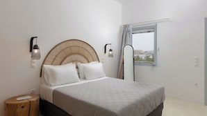 A bedroom with a bed, a round woven headboard, two wall-mounted lights, a window with a view, and a wooden stool.