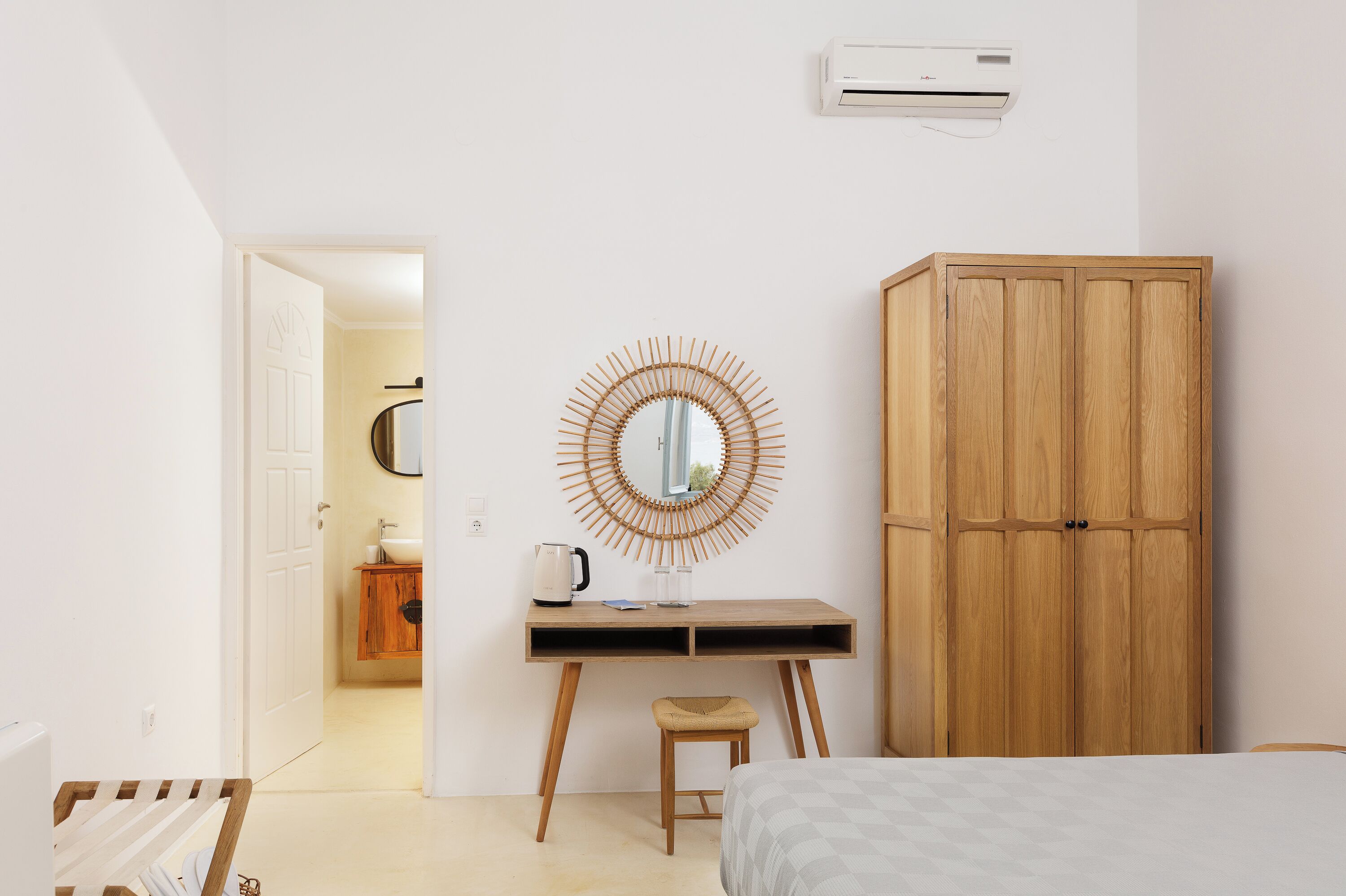 A bedroom with a bed, a wooden wardrobe, a desk with a mirror, and a bathroom visible through an open door.