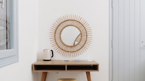 A room with a wooden desk, a woven stool, a wall-mounted circular bamboo decoration, and a white door.